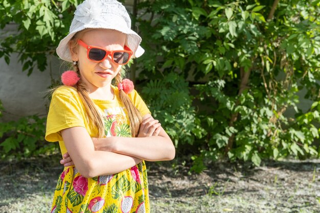 Una bambina emotiva frustrata con un vestito giallo brillante, un cappello bianco e occhiali da sole arricciò il naso