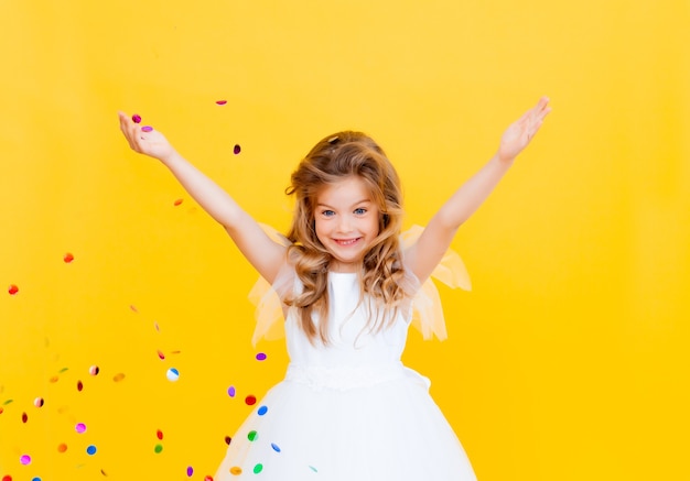 Una bambina felice con i capelli biondi e con un vestito bianco cattura i coriandoli su uno sfondo giallo, concetto di vacanza