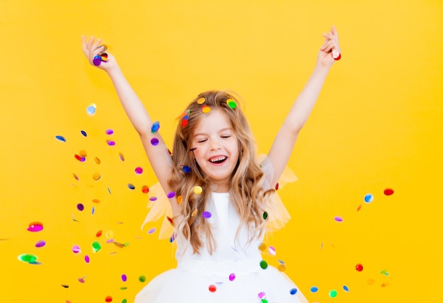 Una bambina felice con i capelli biondi e con un vestito bianco cattura i coriandoli su uno sfondo giallo, concetto di vacanza