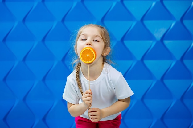 Una bambina felice con un'arancia in mano, guarda la telecamera vestita con una maglietta bianca, isolata su sfondo blu, copia spazio,
