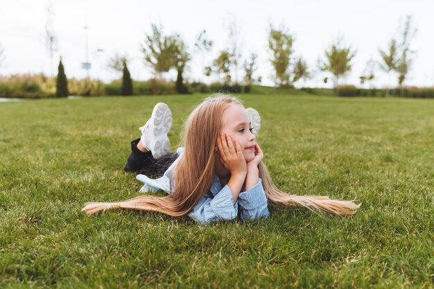 Una bambina felice con una giacca di jeans giace sul prato verde dell'erba un bambino sorridente sta riposando sdraiato sull'erba il concetto di un'infanzia spensierata