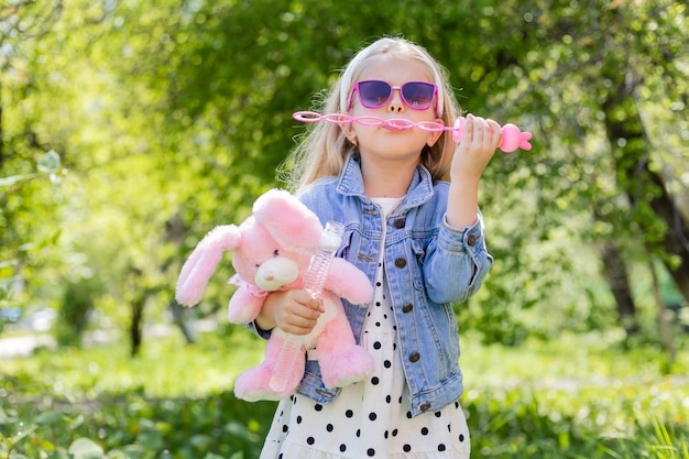 Una bambina felice in estate con gli occhiali da sole gonfia le bolle di sapone tiene un giocattolo nelle sue mani