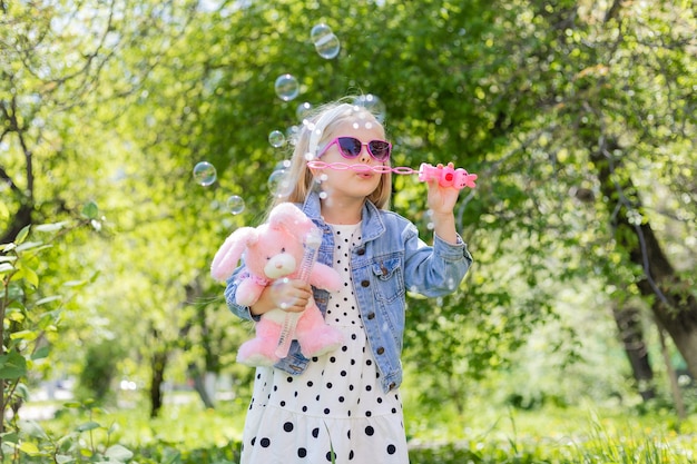 Una bambina felice in estate con gli occhiali da sole gonfia le bolle di sapone tiene un giocattolo nelle sue mani