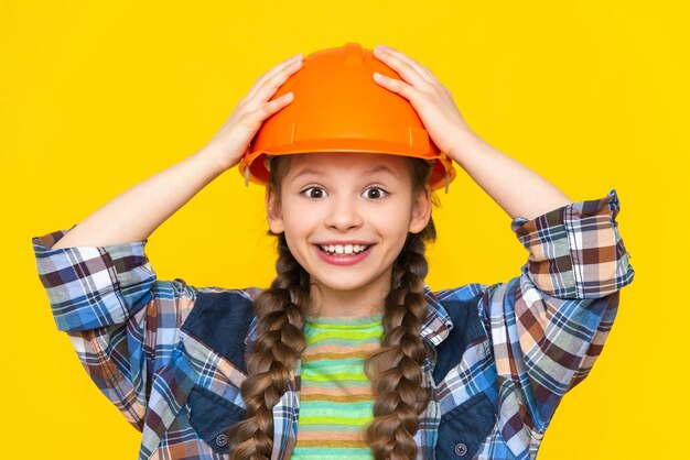 Una bambina felice in un casco da costruzione e una camicia su uno sfondo giallo isolato Il bambino si sta preparando per le riparazioni nella scuola materna La professione di un ingegnere Sfondo giallo isolato