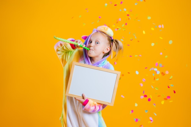 Una bambina felice in un kigurumi unicorno tiene una scheda vuota per il testo su un muro giallo