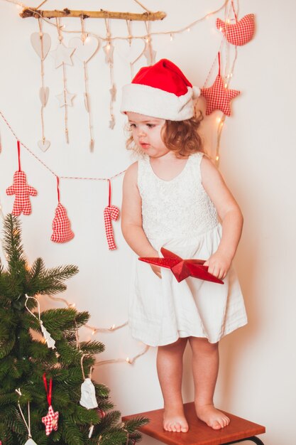 Una bambina graziosa con un berretto di Babbo Natale appende una stella in cima all'albero di Natale