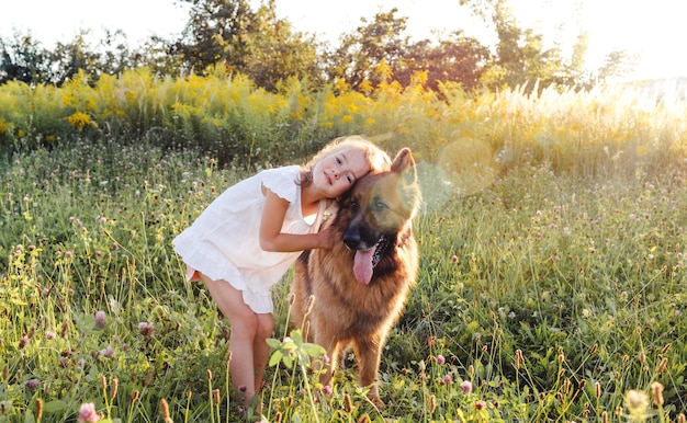 Una bambina in abito bianco abbraccia un grosso cane pastore tedesco in piedi sull'erba verde. Giochi per bambini con un cane.
