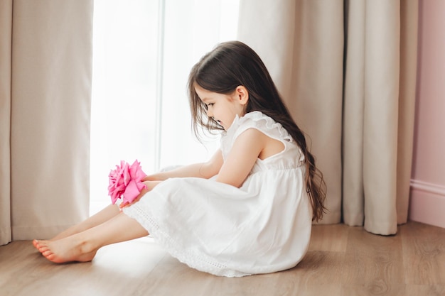 Una bambina in abito bianco è seduta vicino alla finestra e tiene in mano una rosa Un bambino felice alla finestra con un fiore