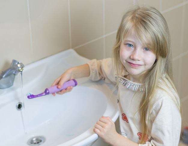 Una bambina in bagno lava uno spazzolino elettrico.