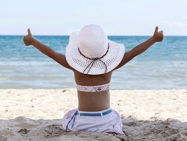 Una bambina in bikini e cappello da spiaggia bianca si siede su una spiaggia sabbiosa con le spalle alla telecamera