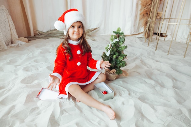 Una bambina in costume da bagno e un cappello di Babbo Natale sta decorando un albero di Natale e si siede sulla sabbia durante lo spazio della copia delle vacanze di Natale