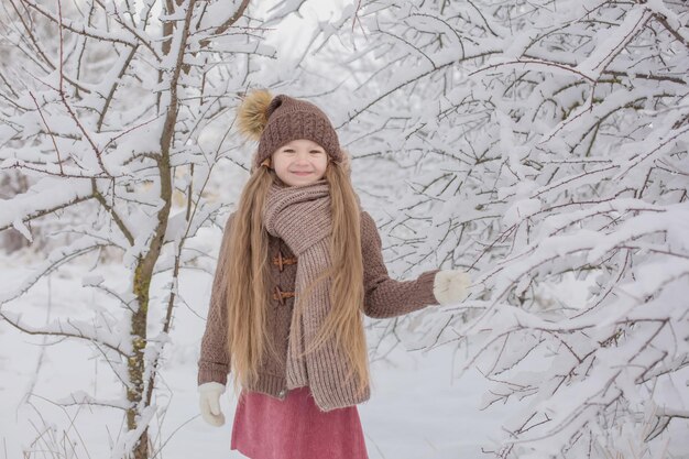 una bambina in maglia marrone cose un cappello e una sciarpa si trova in un parco innevato