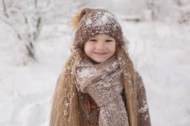 una bambina in maglia marrone cose un cappello e una sciarpa si trova in un parco innevato