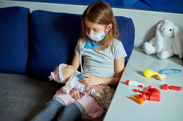 Una bambina in maschera medica si siede sul divano, interpreta un dottore, cura e ascolta la bambola con uno stetoscopio in maschera medica, a casa durante la quarantena e la pandemia