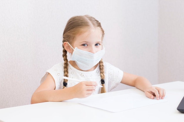 UNA BAMBINA IN MASCHERA SI SIEDE A UN TAVOLO E DISEGNA, SCRIVE. ALUNNO. SCUOLA. IL PRIMO SETTEMBRE. QUARANTENA A CASA