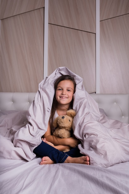 Una bambina in pigiama è seduta sotto una coperta a letto con un orsacchiotto