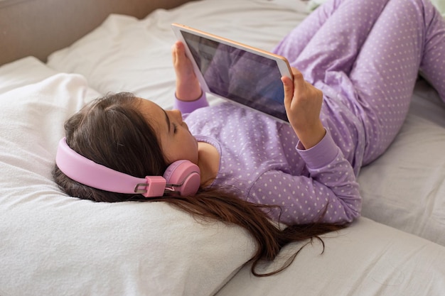 Una bambina in pigiama viola giace su un letto bianco sulla schiena tiene in mano una tavoletta digitale rosa