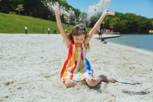 Una bambina in un abito estivo getta sabbia sulla spiaggia per le vacanze estive