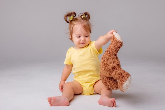 Una bambina in un body giallo si siede e gioca con un orsacchiotto su uno sfondo bianco