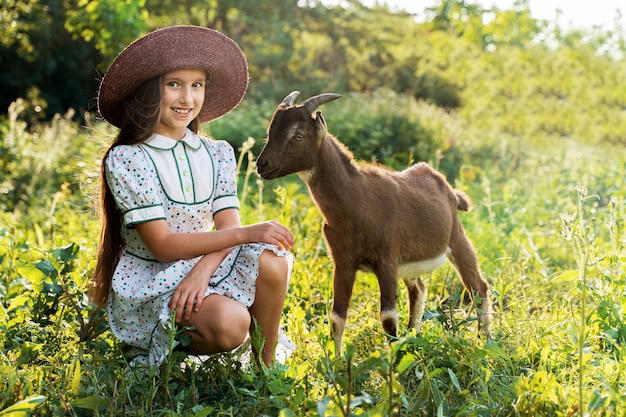 Una bambina in un cappello sta curando una capra in un prato verde