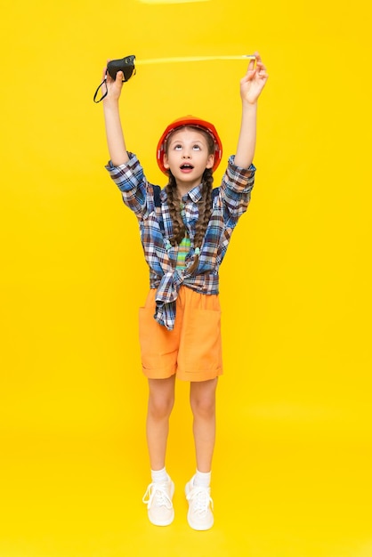 Una bambina in un casco da costruzione arancione tiene un nastro di misurazione Il concetto di rinnovamento nella stanza dei bambini Il bambino è un ingegnere Verticale A tutta altezza Sfondo giallo isolato