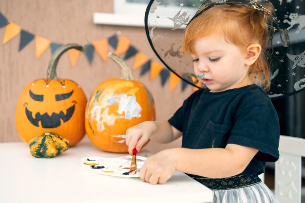 Una bambina in un costume da strega dipinge zucche per halloween a casa il concetto di crea...