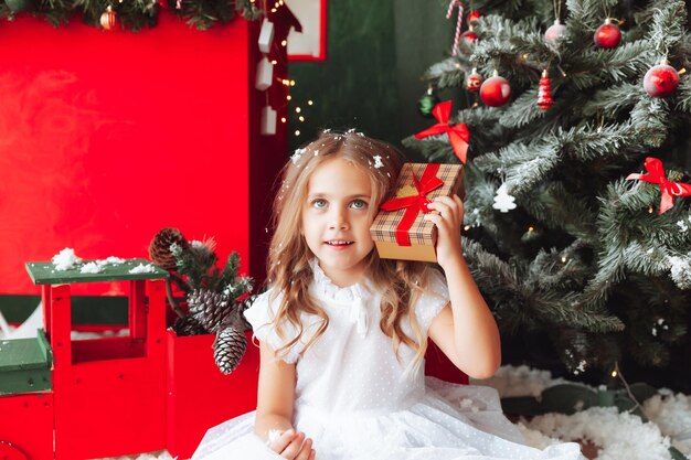 Una bambina in un vestito bianco è seduta sul pavimento e tiene in mano un regalo di Capodanno sullo sfondo di un albero di Natale il concetto di Natale e Capodanno