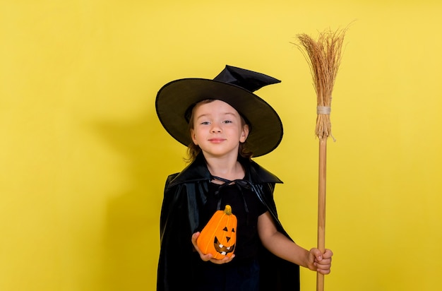 Una bambina in un vestito streghe con un cappello con una zucca e una scopa su una parete isolata gialla con spazio per il testo