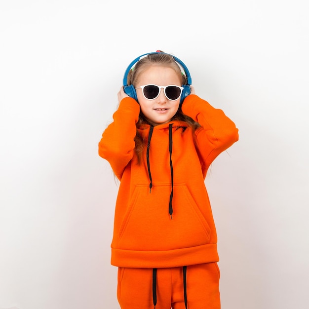 Una bambina in una felpa con cappuccio arancione occhiali da sole e cuffie blu ascolta musica Ritratto su uno sfondo bianco