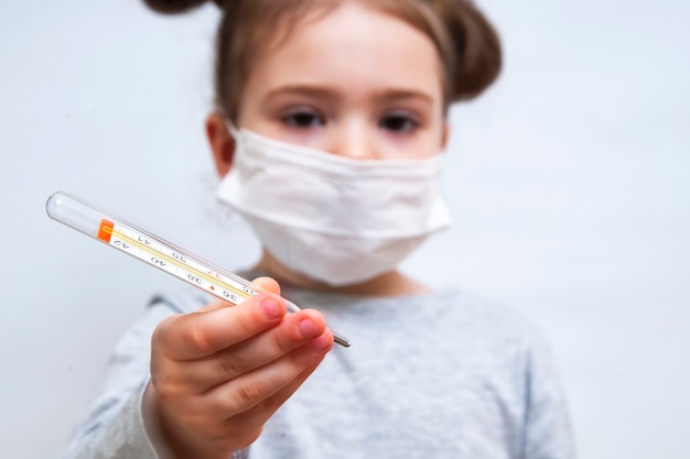 Una bambina in una maschera medica mostra il termometro. Protezione dell'epidemia di coronavirus