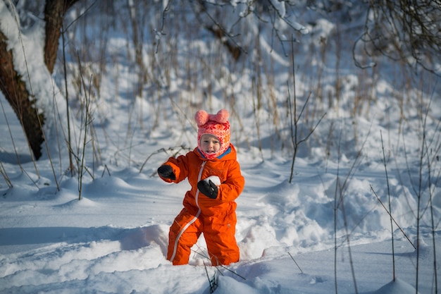 Una bambina in una tuta arancione gioca a palle di neve in inverno nella foresta.