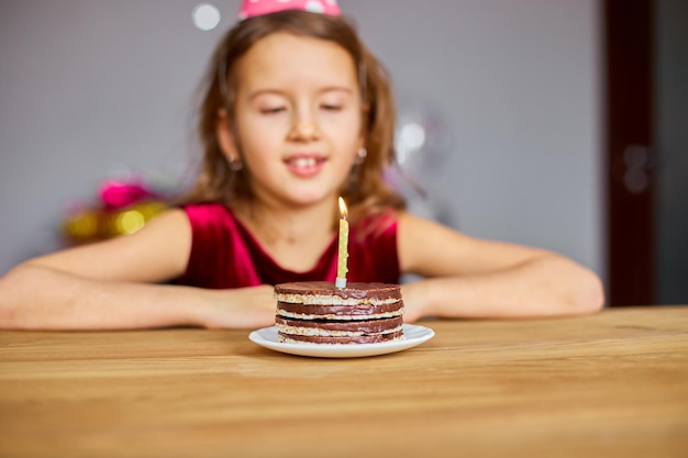 Una bambina indossa un cappello di compleanno esprime un desiderio guardando una torta di compleanno con candele accese per un concetto di festa