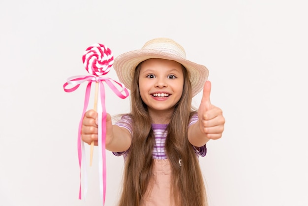 Una bambina mostra un lecca-lecca e sorride Un bambino con un prendisole e un cappello estivo su uno sfondo bianco isolato mostra un pollice in alto