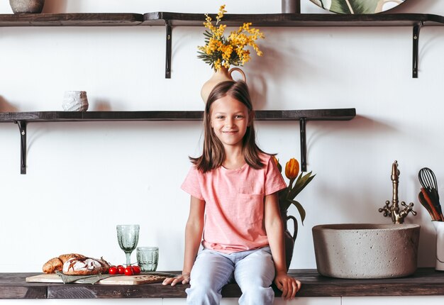 Una bambina positiva si siede sul tavolo della cucina. Guarda la telecamera