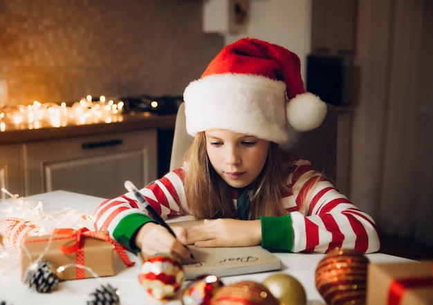 Una bambina prima di Natale scrive una lettera a Babbo Natale sul tavolo con ghirlande. bambino scrive una lettera a babbo natale.