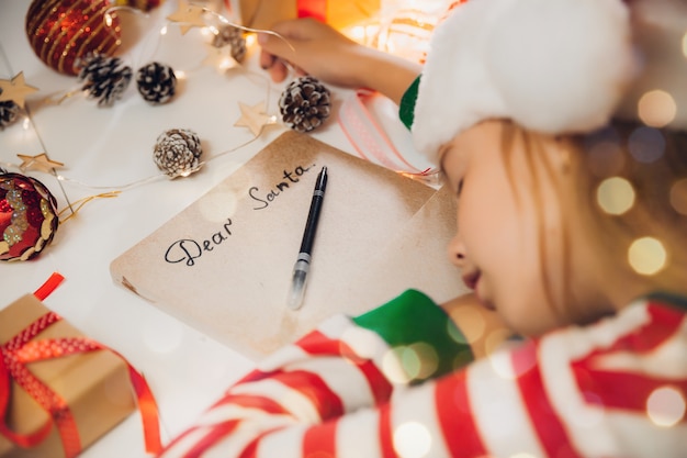 Una bambina prima di Natale scrive una lettera a Babbo Natale sul tavolo con ghirlande. bambino scrive una lettera a babbo natale.