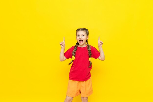 Una bambina punta il dito contro uno spazio vuoto per la tua pubblicità su uno sfondo giallo isolato Un bel bambino sorride in una maglietta rossa con due treccine Copia spazio