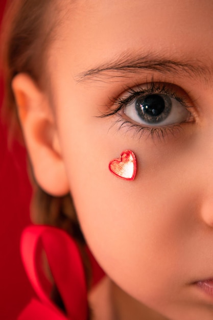 Una bambina seria guarda nella fotocamera con il cuore sulla guancia con le trecce su sfondo rosso Foto di alta qualità