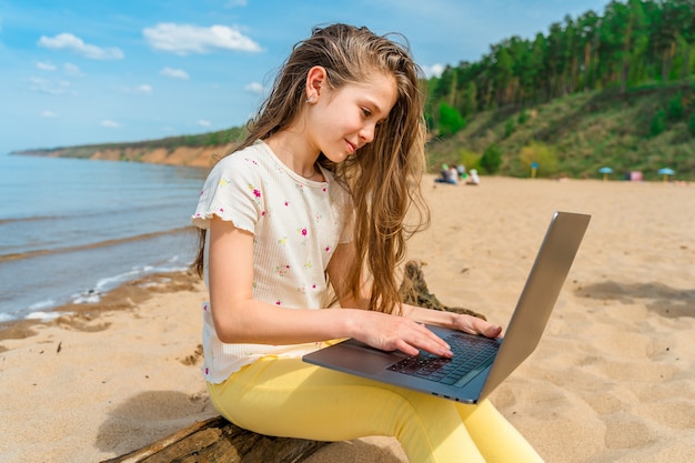 Una bambina si siede su un tronco sulla spiaggia con un laptop in grembo Il bambino usa un laptop