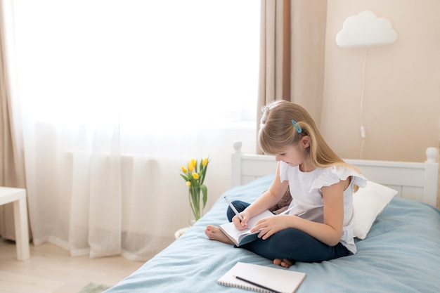 Una bambina si siede sul letto in camera da letto e scrive in un libro blu. Concetto di educazione. Istruzione domiciliare. Compiti a casa.