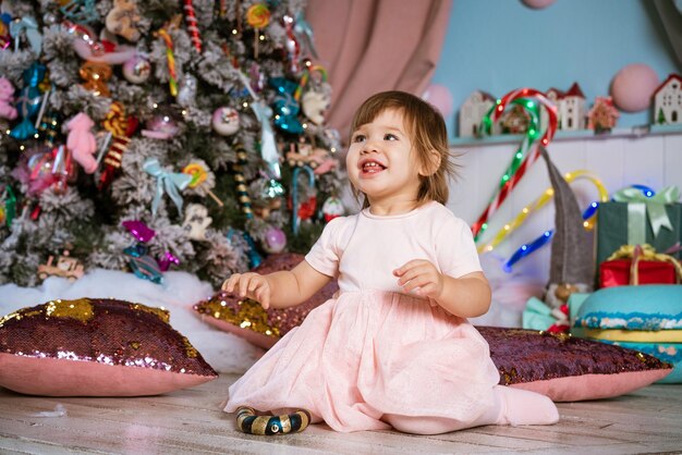 Una bambina si siede sul pavimento con regali accanto all'albero di Natale in abito rosa, felice bambino caucasico è...
