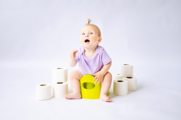 Una bambina sorridente carina è seduta su una pentola con rotoli di carta igienica isolati su sfondo bianco
