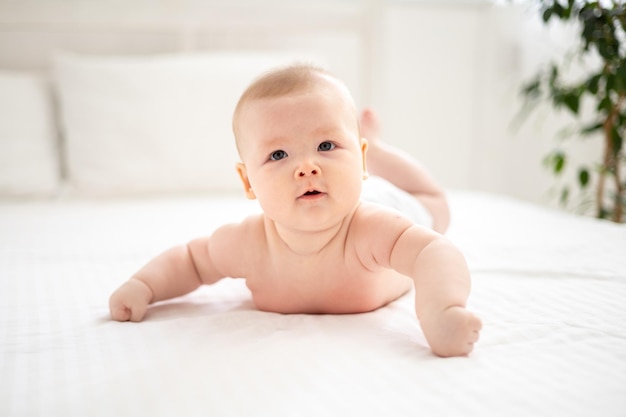 Una bambina sorridente carina in mutandine bianche è sdraiata sul letto nella camera da letto della casa su lenzuola bianche sullo stomaco guardando la telecamera sorridente bambino felice