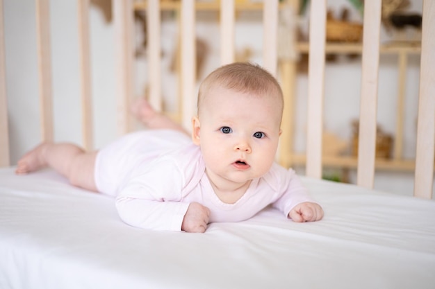 Una bambina sorridente carina in un body è sdraiata in una culla a casa su lenzuola bianche sullo stomaco guardando la telecamera sorridente bambino felice