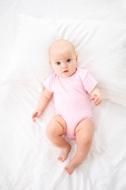 Una bambina sorridente carina in un body rosa è sdraiata sul letto nella camera da letto della casa su lenzuola bianche sulla schiena guardando la telecamera sorridente bambino felice