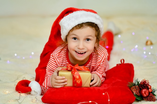Una bambina sorridente con il cappello rosso di Babbo Natale e gli abiti rossi di Natale giace sul letto con un regalo. Preparazione per la celebrazione. Capodanno. il Natale dei bambini. Atmosfera. Casa.