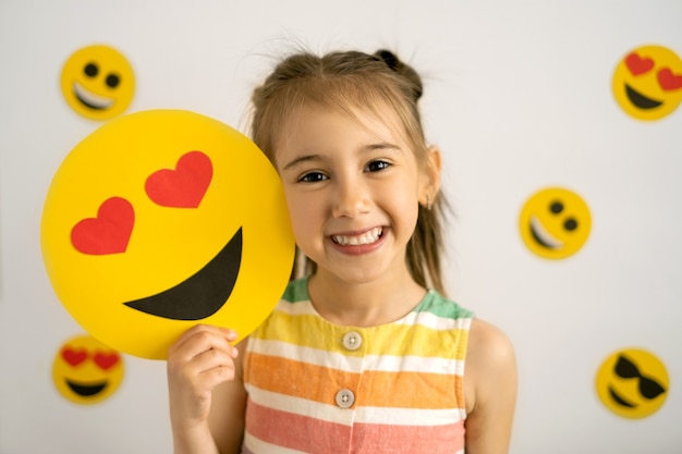 Una bambina sorridente con tutti i denti tiene in mano un'emoji di cartone dell'amore