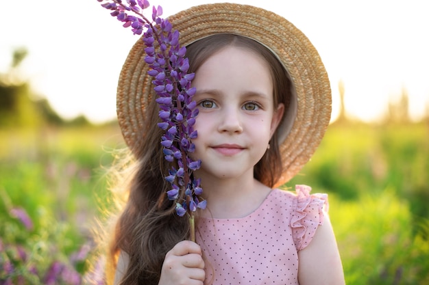 Una bambina sorridente con un cappello di paglia tiene un fiore di lupino Ragazza in un campo di lupini Vacanze estive