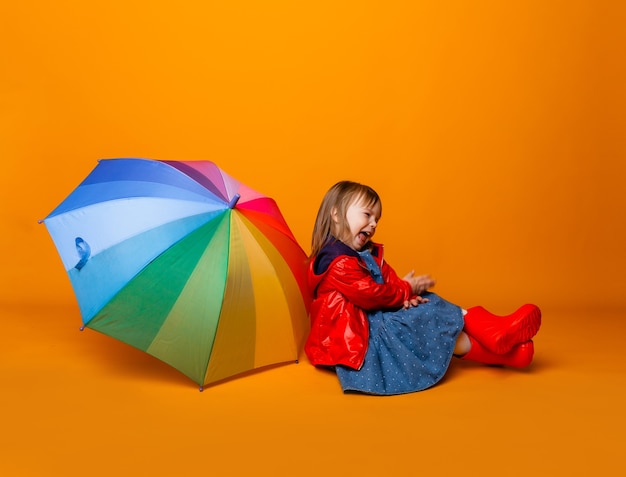 Una bambina sorridente con una giacca rossa e stivali, con in mano un ombrello, si erge su uno sfondo giallo. bambina in abiti luminosi tiene un ombrello arcobaleno.