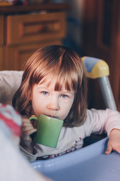 Una bambina sorridente di un anno si siede a un tavolo per bambini su un seggiolone e mangia con un cucchiaio da una ciotola. Sfondo colorato. Mangiare sano per i bambini. Cibo per neonato.